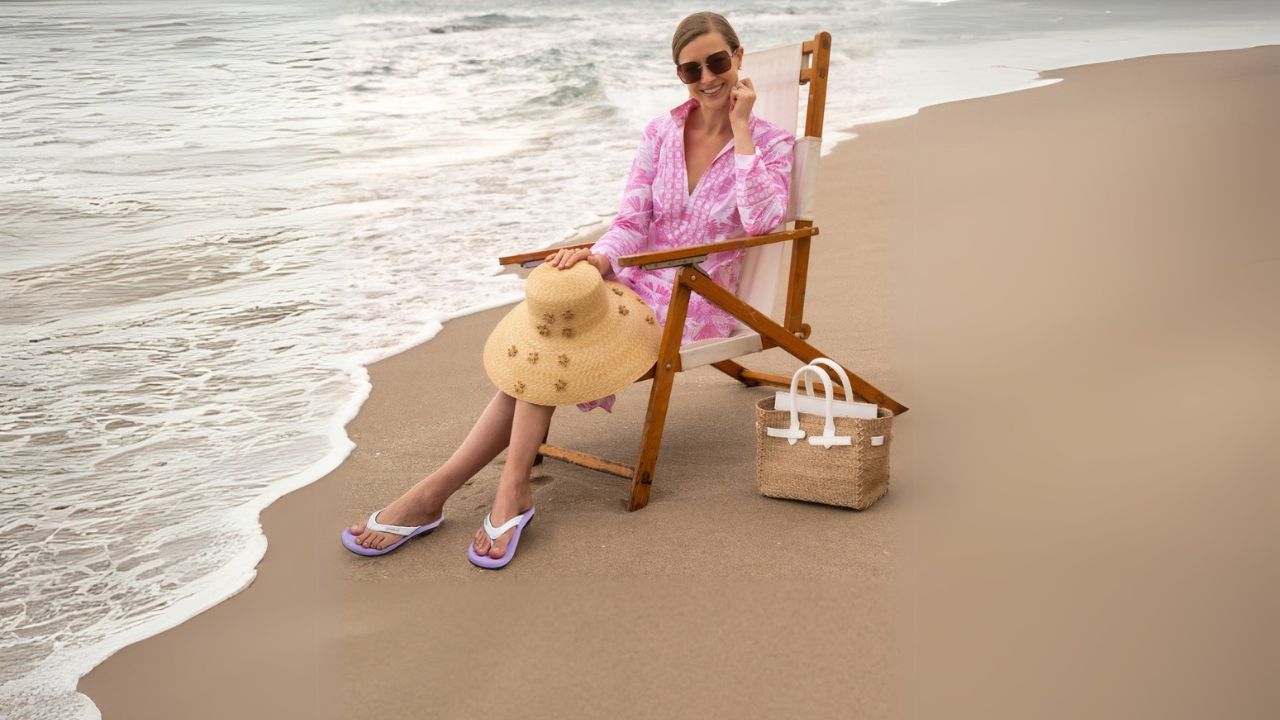 A woman in a pink dress sits in a chair on the beach, with waves gently lapping at the shore in the background.