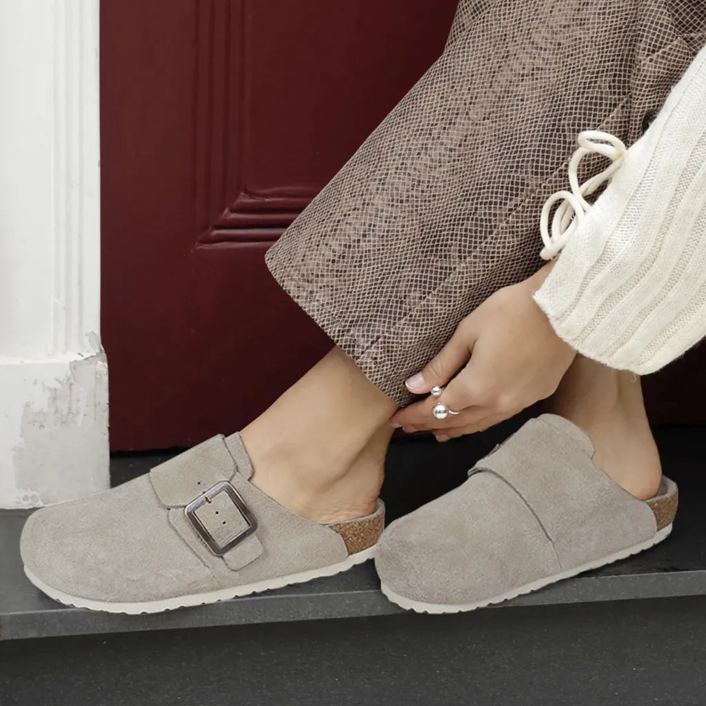 Person wearing gray suede slippers with a buckle, sitting on a step.
womens clogs
